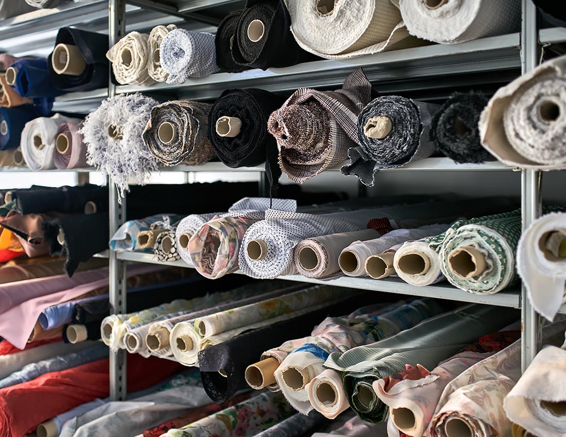 Textured multicolored fabric rolls are lying on gray shelves inside a warehouse. They are different sizes. Closeup horizontal photo with selective focus.